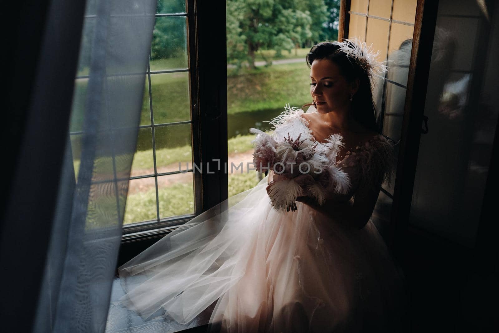 A bride in a wedding dress and a bouquet sits at an open old window and looks by Lobachad