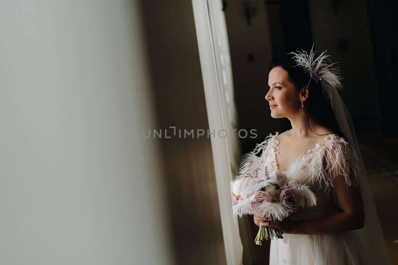 The bride in a wedding dress and with a bouquet stands at the old window and looks by Lobachad