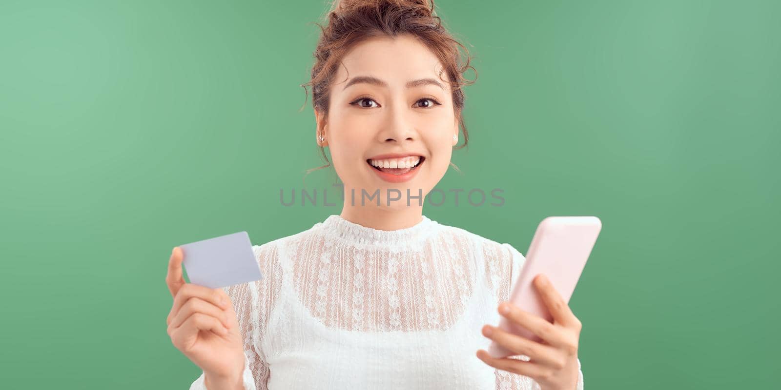 Portrait of a happy young girl with long brunette hair standing over green background, holding mobile phone, showing plastic credit card