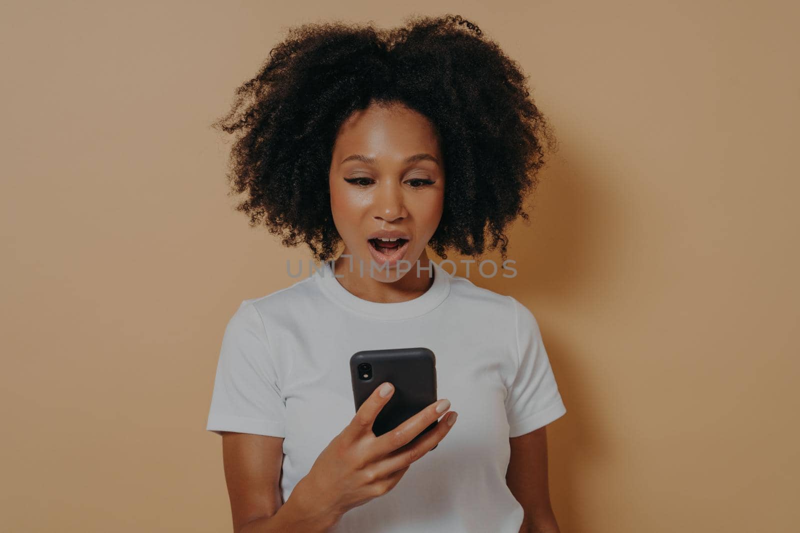 Surprised young african female looking at smartphone with shocked face expression, reading unexpected news in internet or chatting with friend, dressed in white tshirt, isolated over beige background