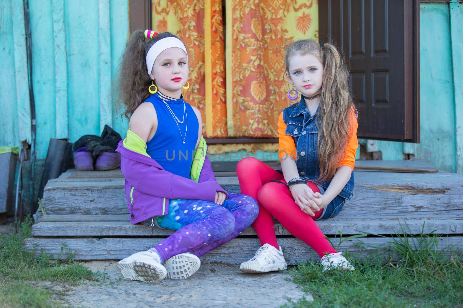 Two funny little girls with bright makeup dressed in the style of the nineties are sitting on the village porch of the house. Russian village children.