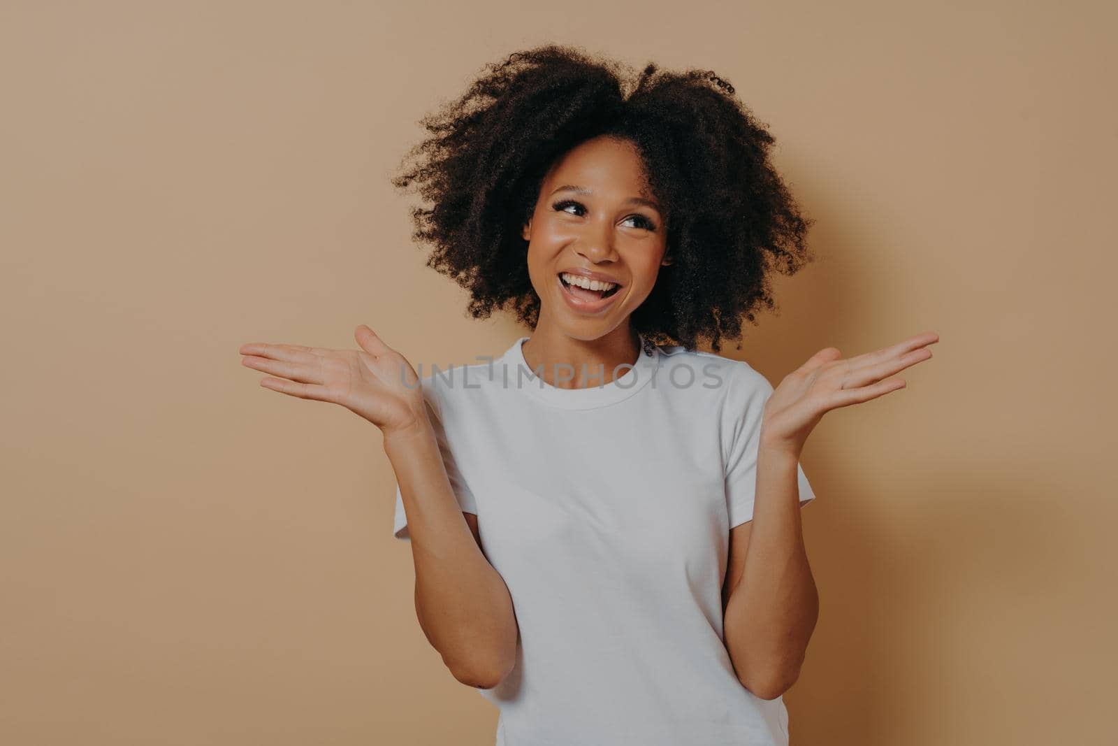 Young smiling mixed race woman raising hands with hesitation, isolated on beige background by vkstock