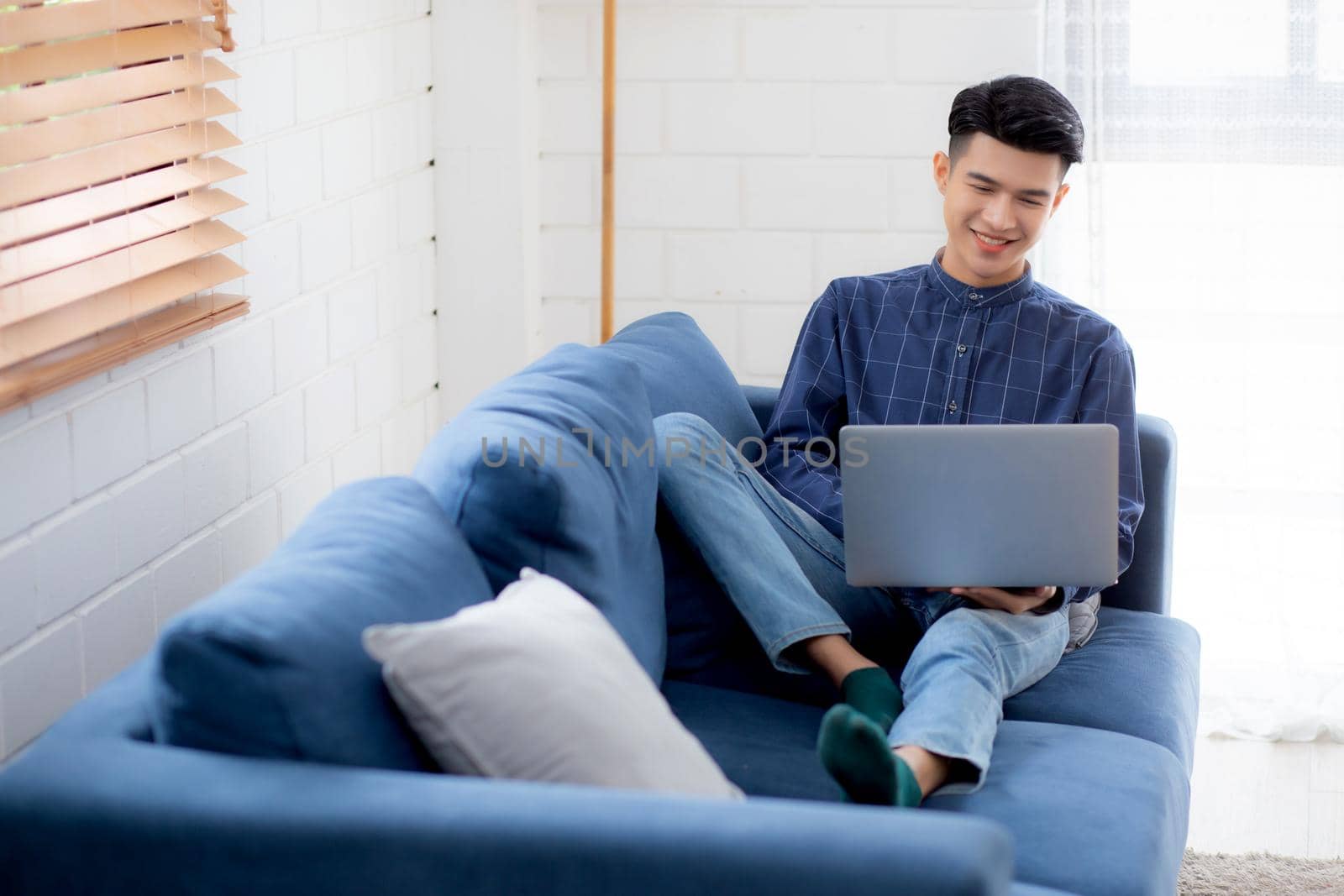 Young asian businessman smile and work from home with laptop computer online to internet on sofa in living room, freelance man using notebook on couch with comfort, new normal, lifestyle concept.