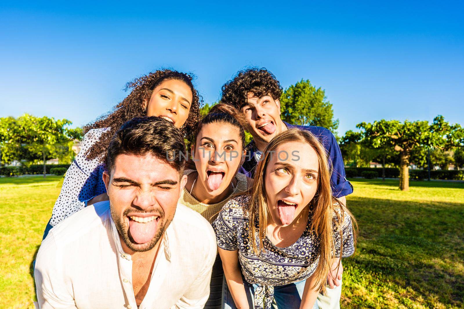 Group of silly young multiracial millennial friends making funny faces with tongue, open mouth, and squinting eyes posing for a portrait in city park. Live your life lightly while having fun in nature by robbyfontanesi