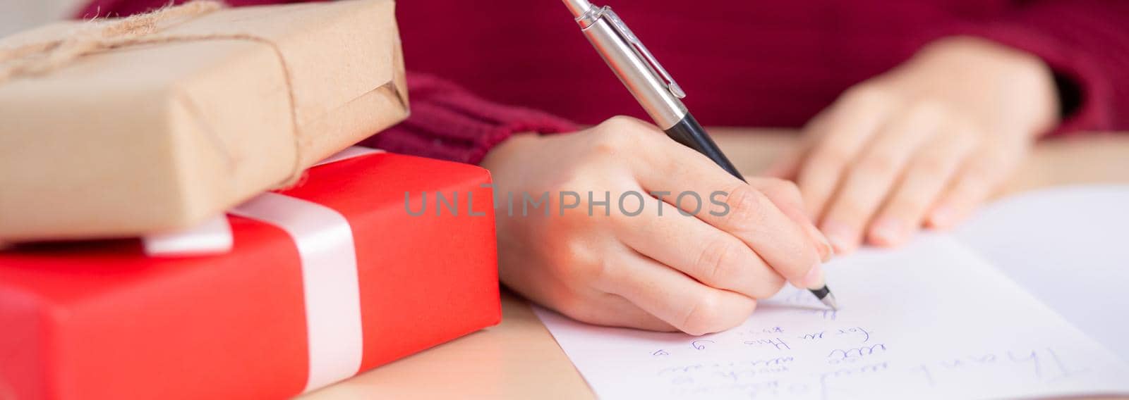 Closeup hand of young asian woman writing postcard in Christmas day at home, eve and celebrate, female writing message on greeting card with giving gift box in holiday, congratulation and celebration.