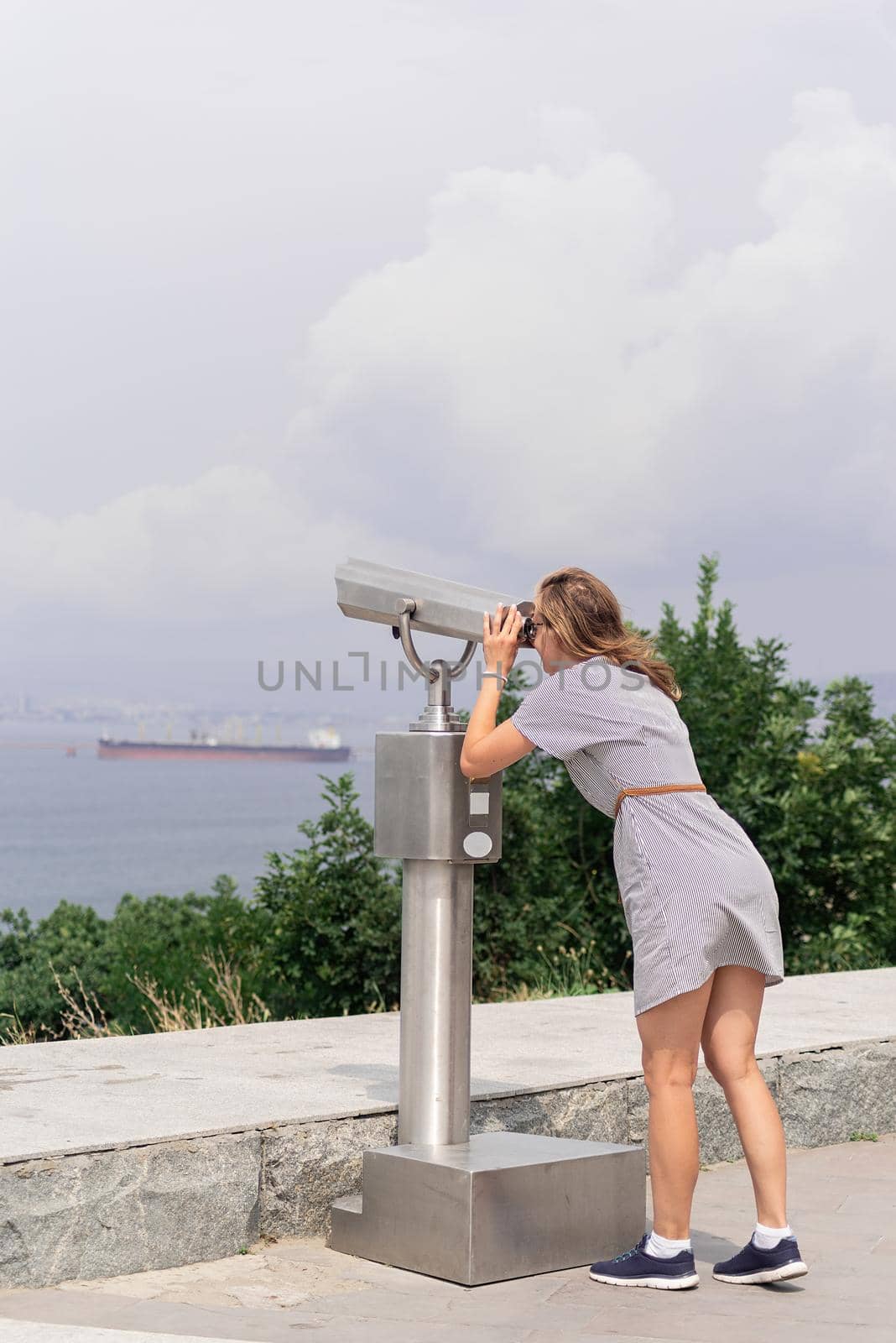 young woman using stationary binoculars on a warm summer day on a hill against the background of sea and city by Desperada