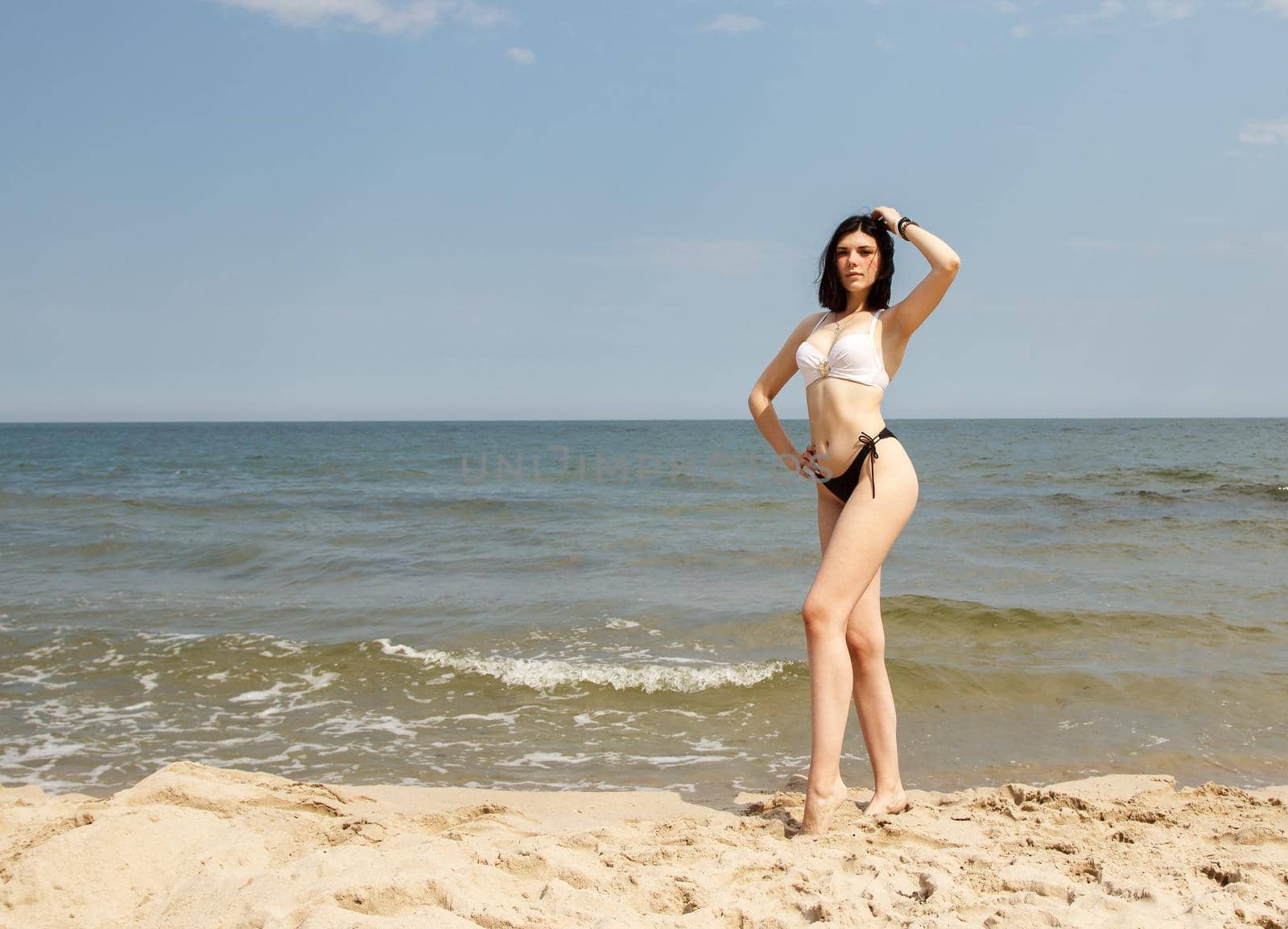 young brunette woman in bikini stands on the sand by the sea on summer day