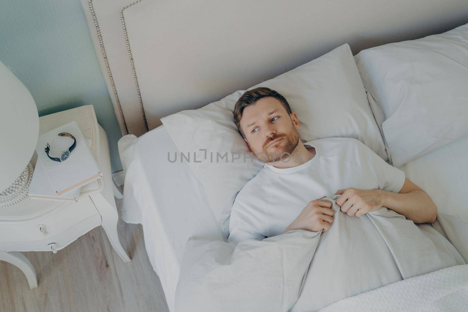 View from above of stressed young caucasian male lying in bed with his eyes open and being unable to sleep, man feeling unhappy and tired from insomnia problems. Bedtime and rest trouble concept