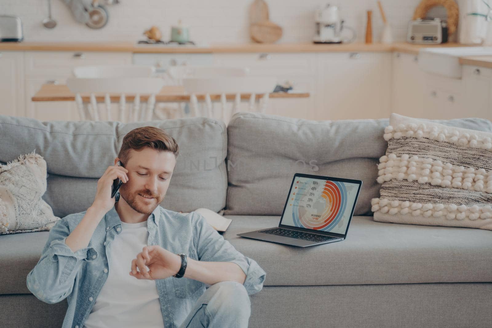 Young company male worker calling his supervisor while uploading infographics and sending them to his boss, checking time hoping he isn't late, sitting on floor while resting against couch