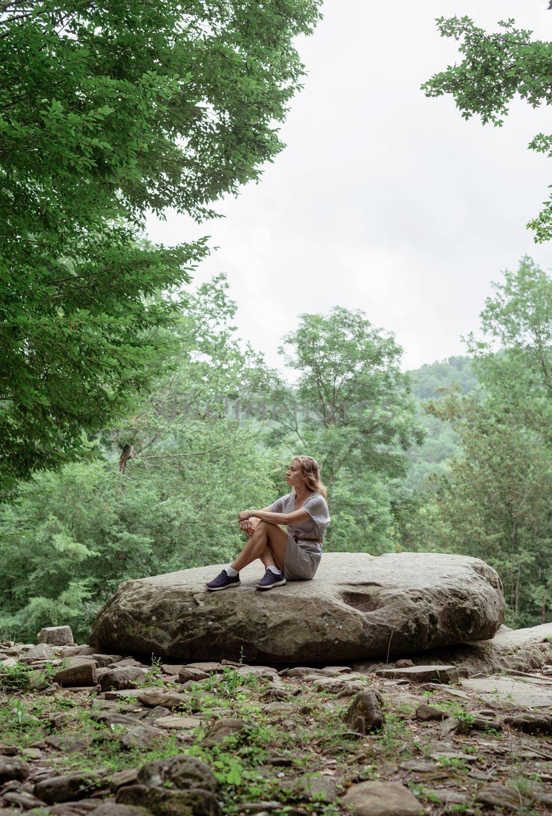 Travel concept. Young woman in summer dress sitting on a big rock in the forest, having rest or meditating