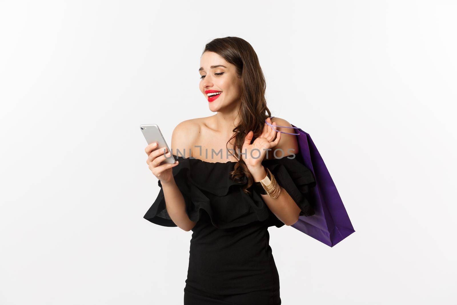 Beauty and shopping concept. Gorgeous woman in elegant black dress and makeup, order online on smartphone, holding bag and smiling, standing over white background by Benzoix