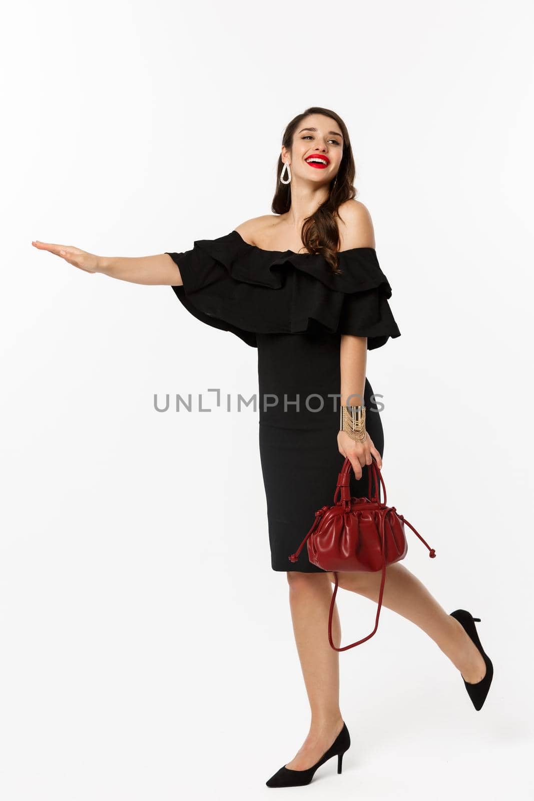 Beauty and fashion concept. Full length of glamour woman in black dress and high heels raising hand to stop taxi, need a ride, standing over white background.