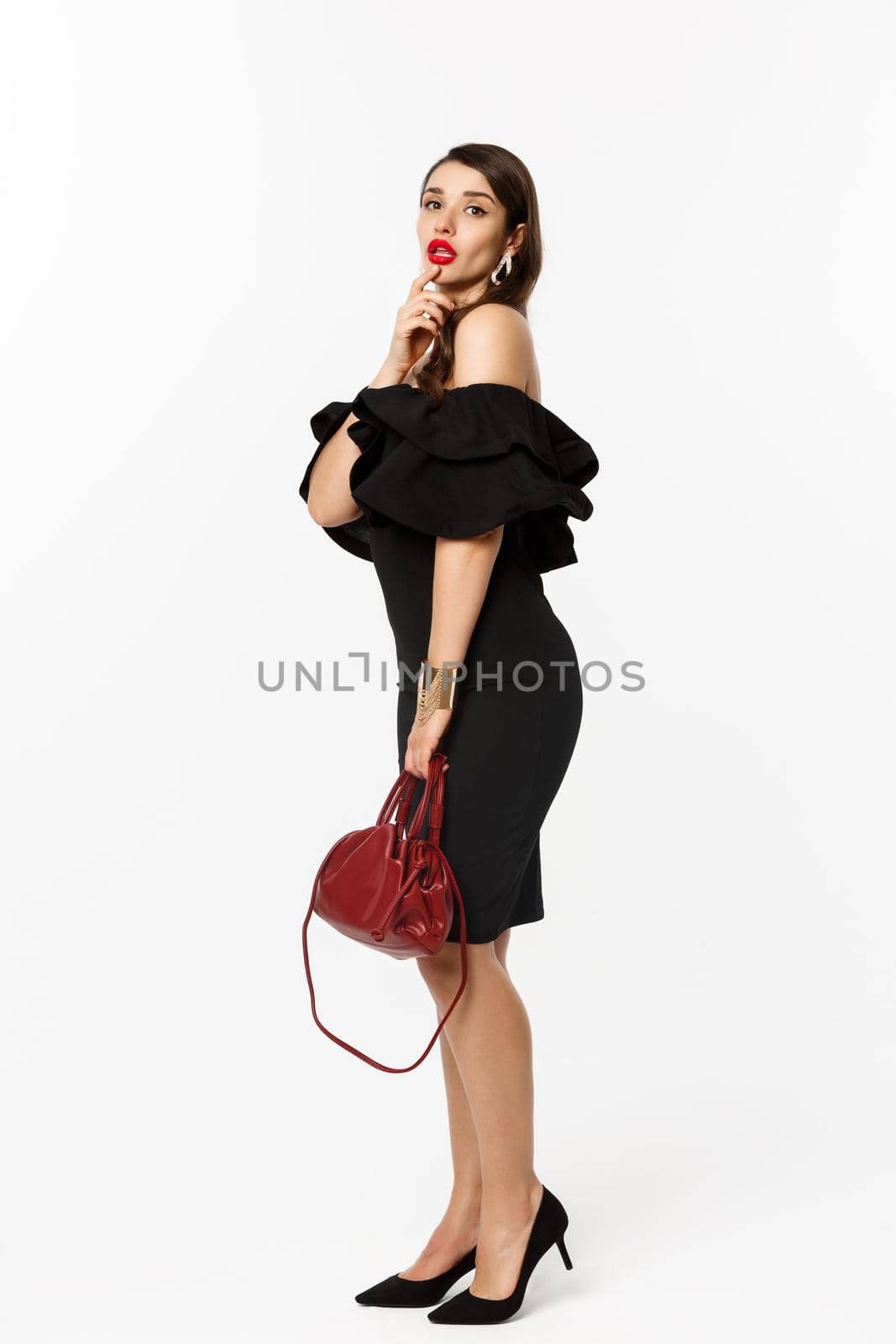 Beauty and fashion concept. Full length of sensual young woman in black elegant dress, high heels and purse, touching lip flirty and gazing at camera, white background by Benzoix