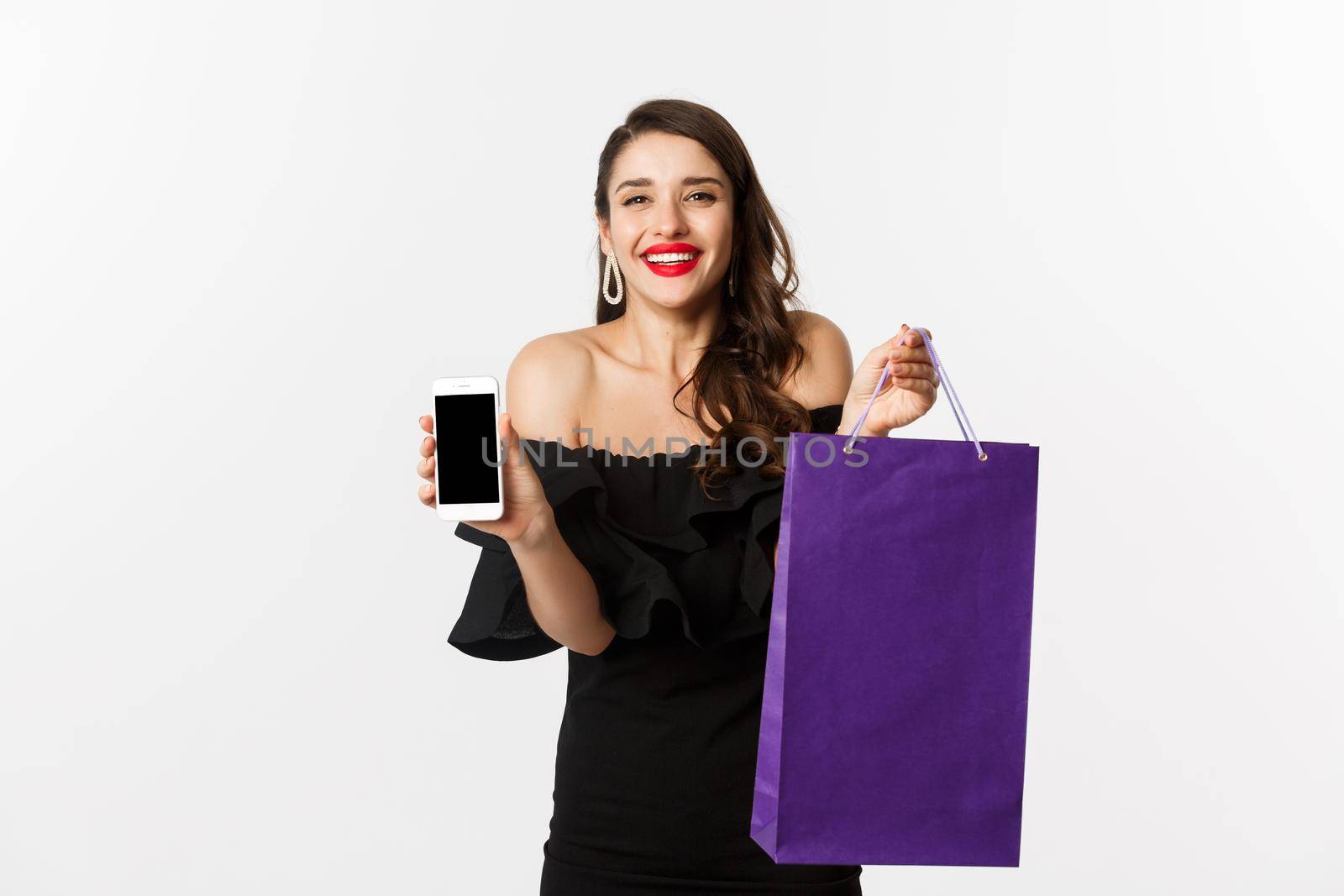 Beauty and shopping concept. Beautiful and stylish woman showing mobile phone screen and bag, buying online, standing over white background.