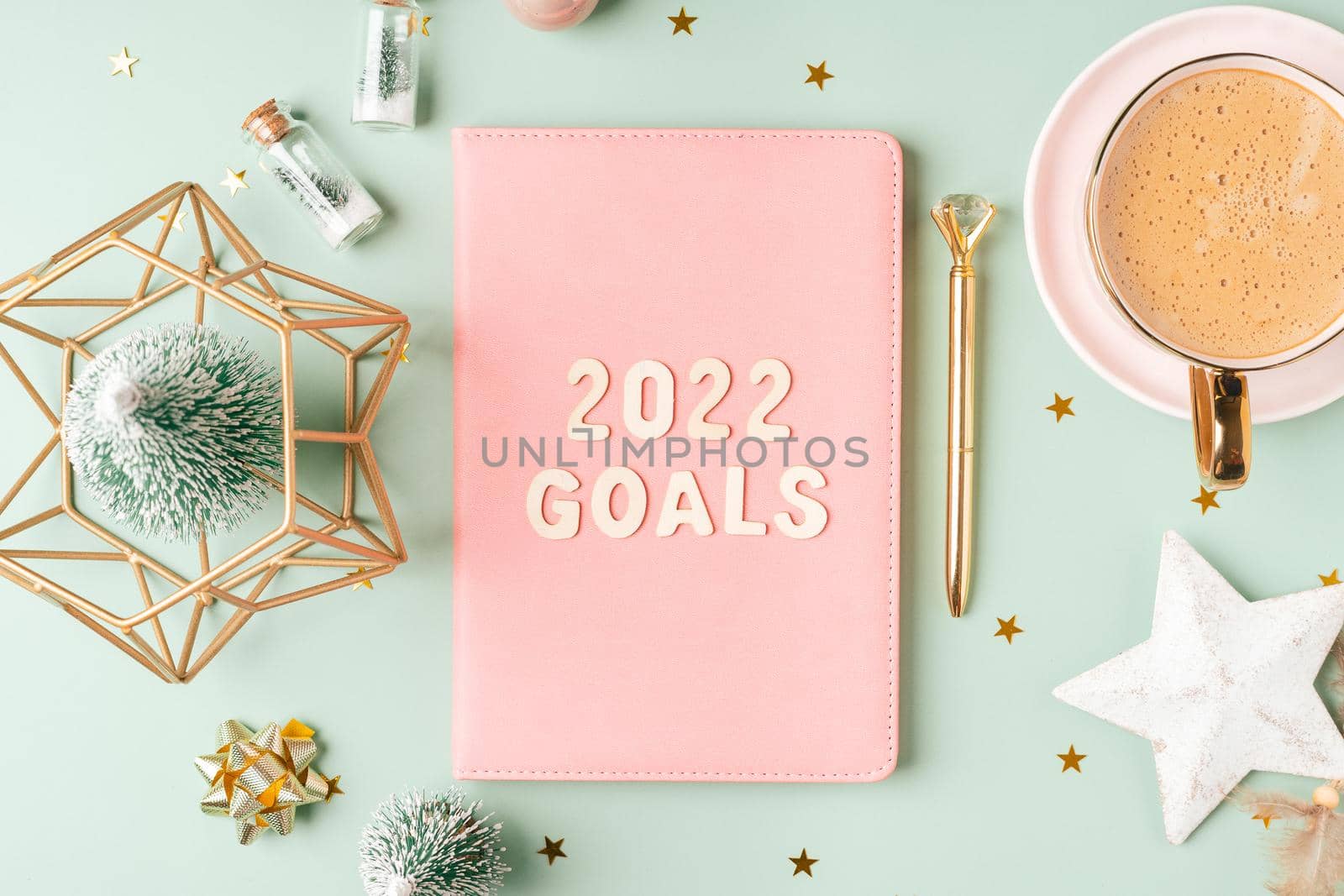 Top view Desktop Christmas pink notepad with 2022 goals letters text. Flat lay on green mint table background with planner, cup of coffee, candle, Christmas decoration, notebook and stationery by Ostanina