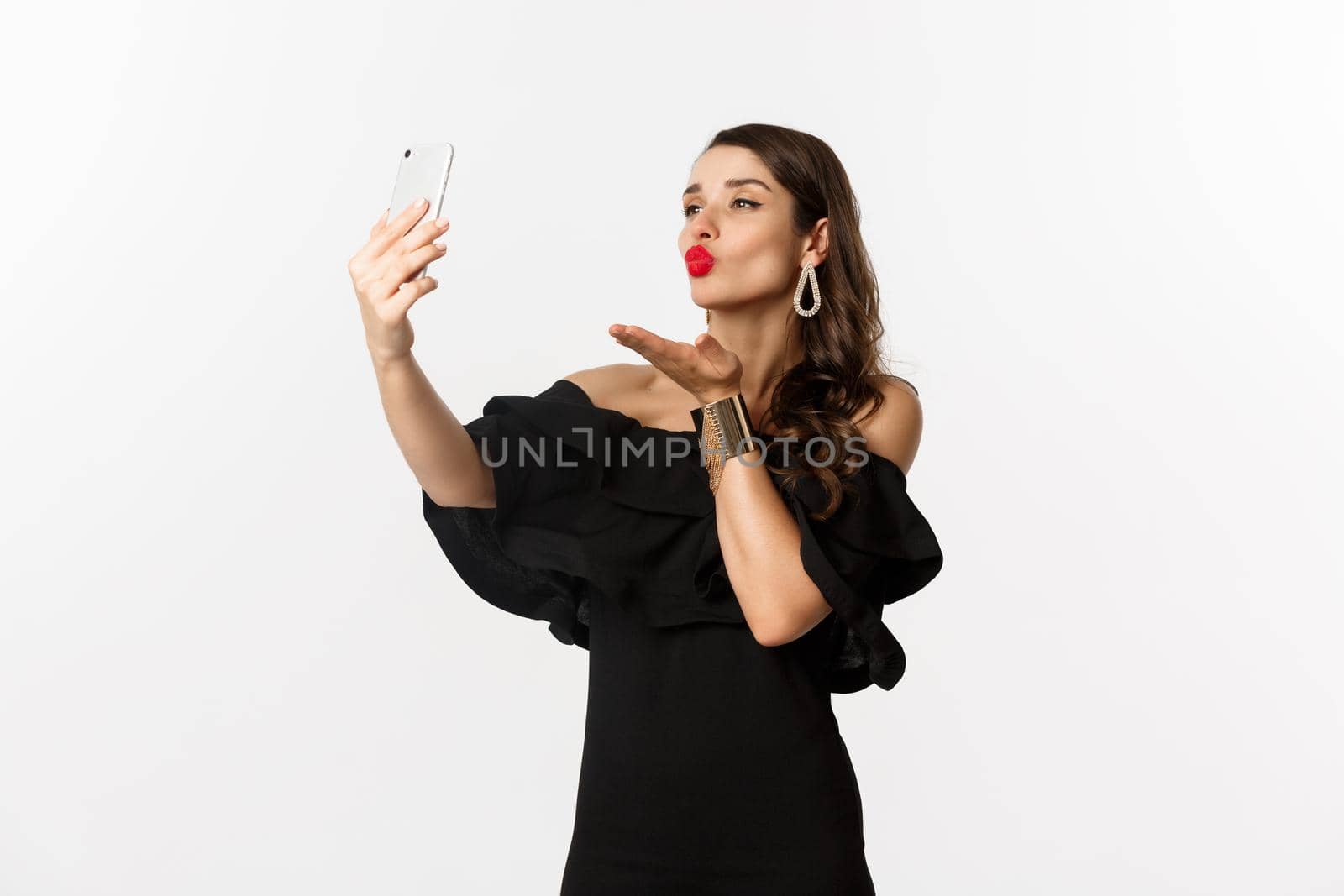 Stylish young woman in black dress, partying and taking selfie on mobile phone, sending air kiss at smartphone camera, standing over white background.