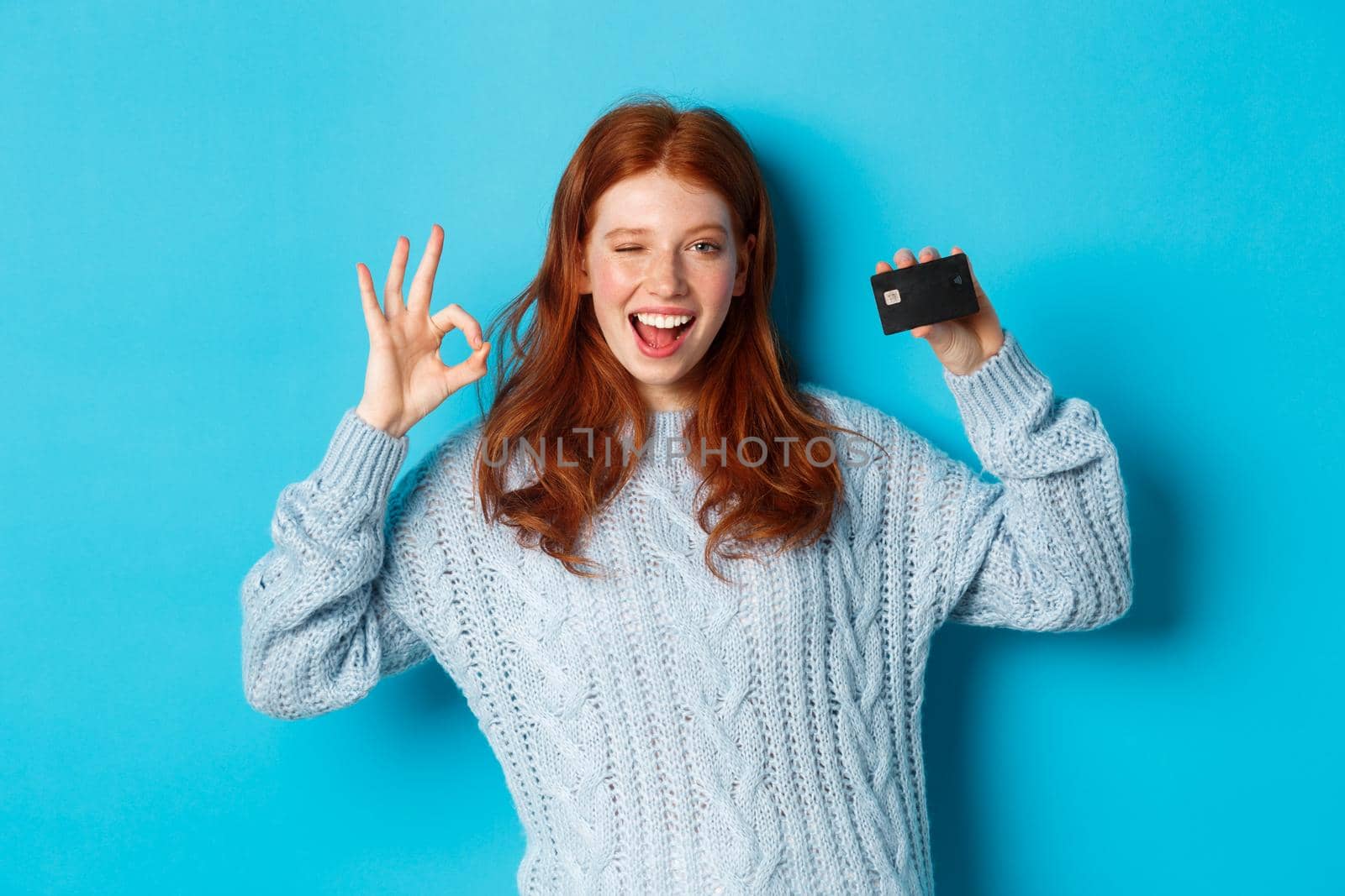 Happy redhead girl in sweater showing credit card and okay sign, recommending bank offer, standing over blue background.