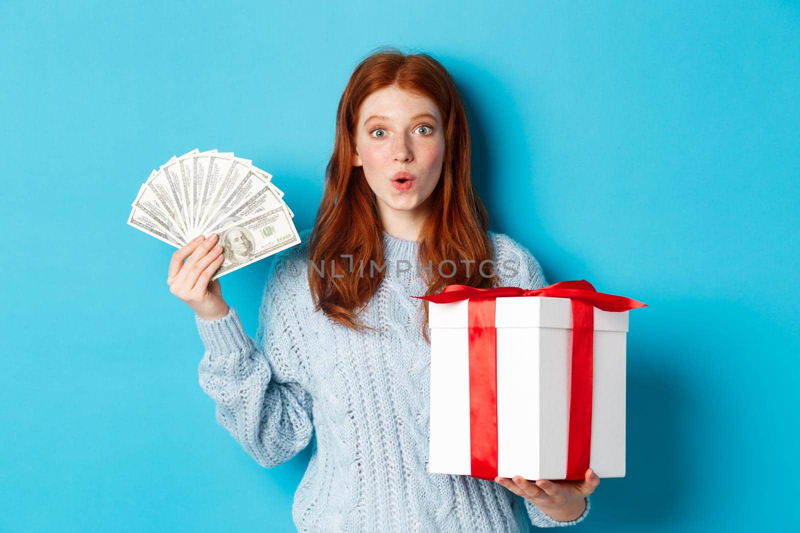 Christmas and shopping concept. Excited redhead girl looking at camera, holding big New Year gift and dollars, buying presents, standing over blue background.