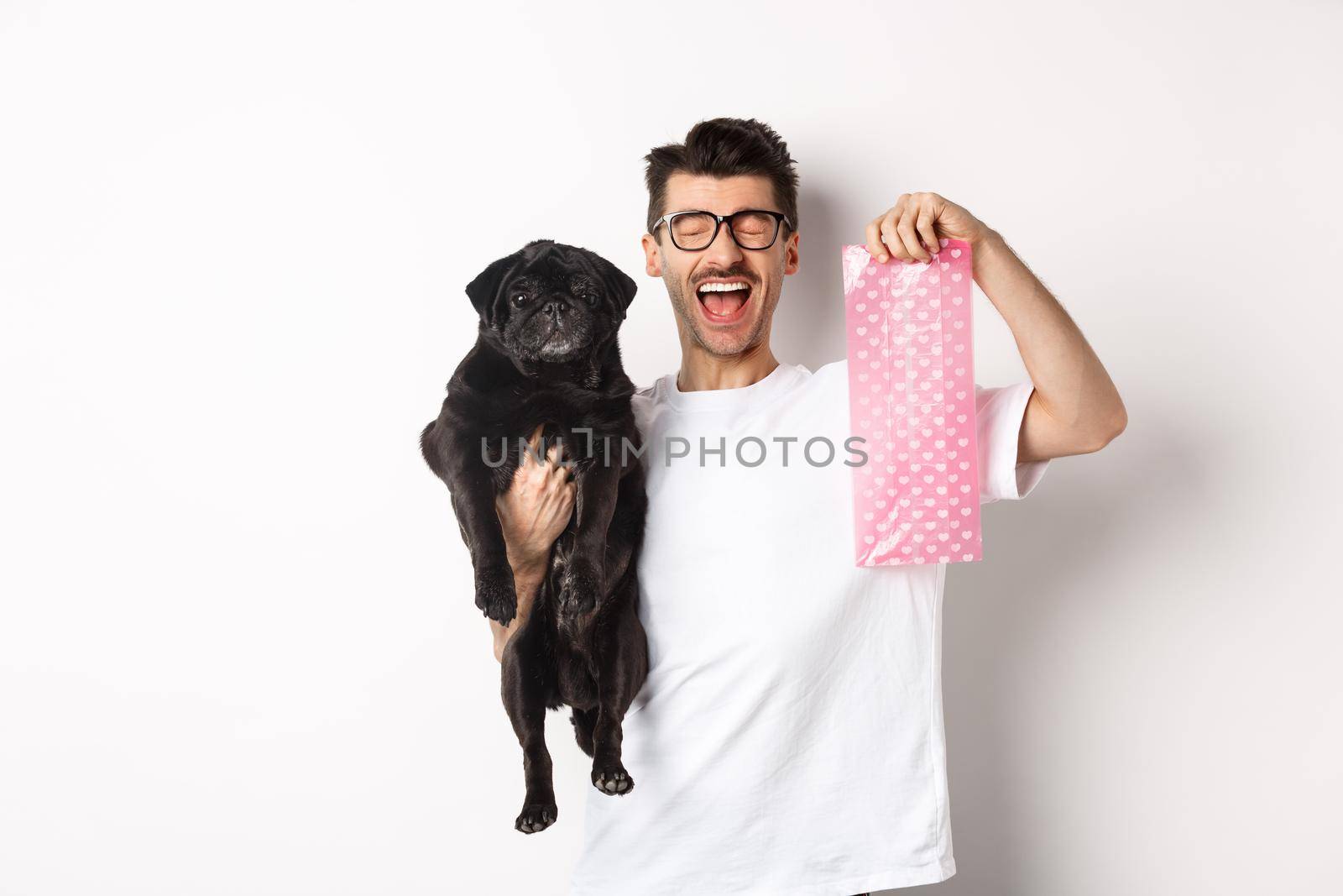 Image of happy man, pet owner, holding cute black pug and dog poop bag, standing over white background.