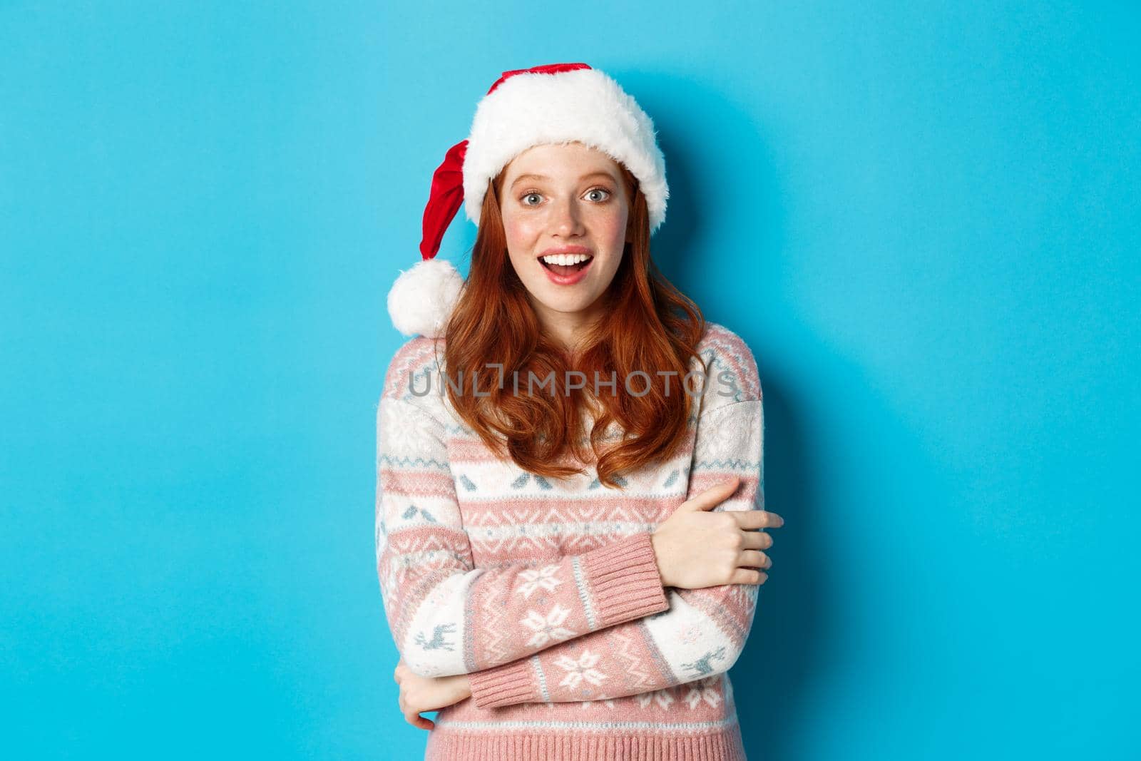 Winter and Christmas Eve concept. Amazed redhead girl in santa hat staring at camera in awe, smiling excited, standing over blue background.