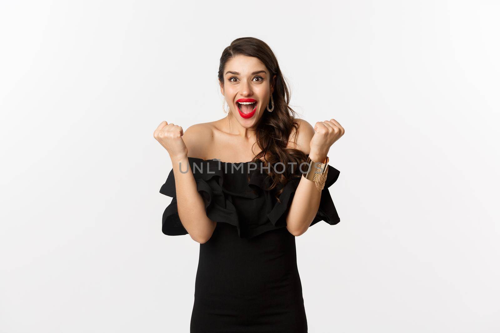 Fashion and beauty. Successful pretty woman in black dress celebrating, rejoicing of winning, triumphing over white background by Benzoix