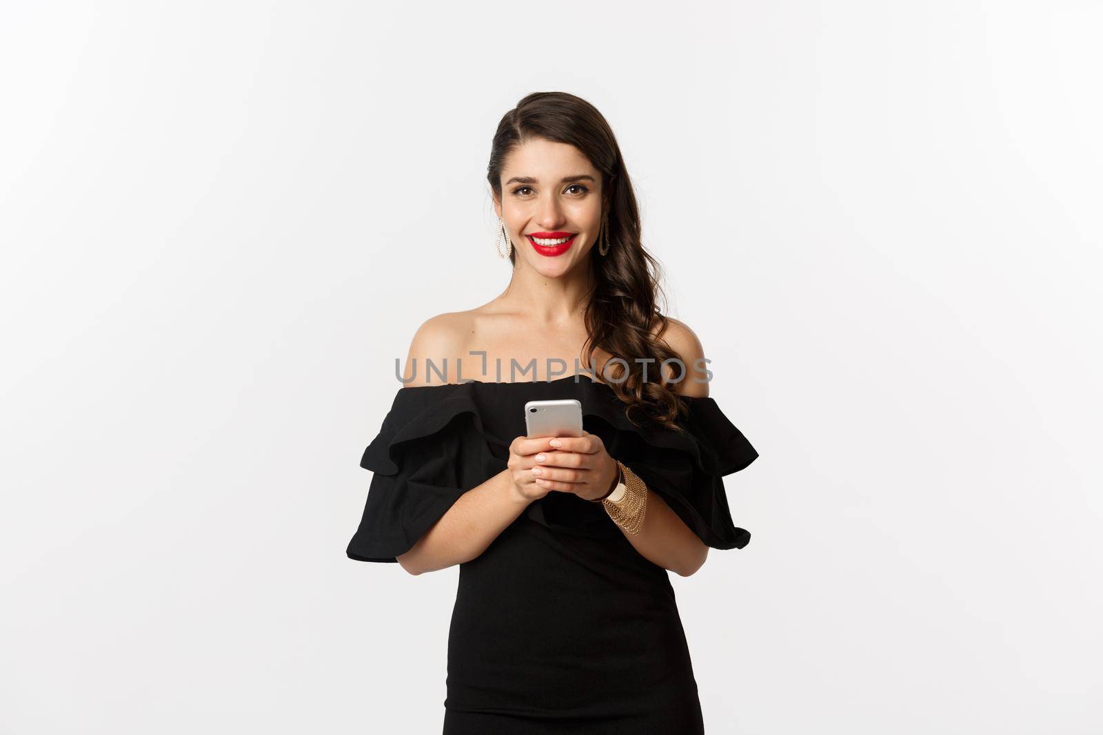 Online shopping concept. Attractive young woman in black dress, reading text message, using mobile phone and smiling, standing over white background by Benzoix