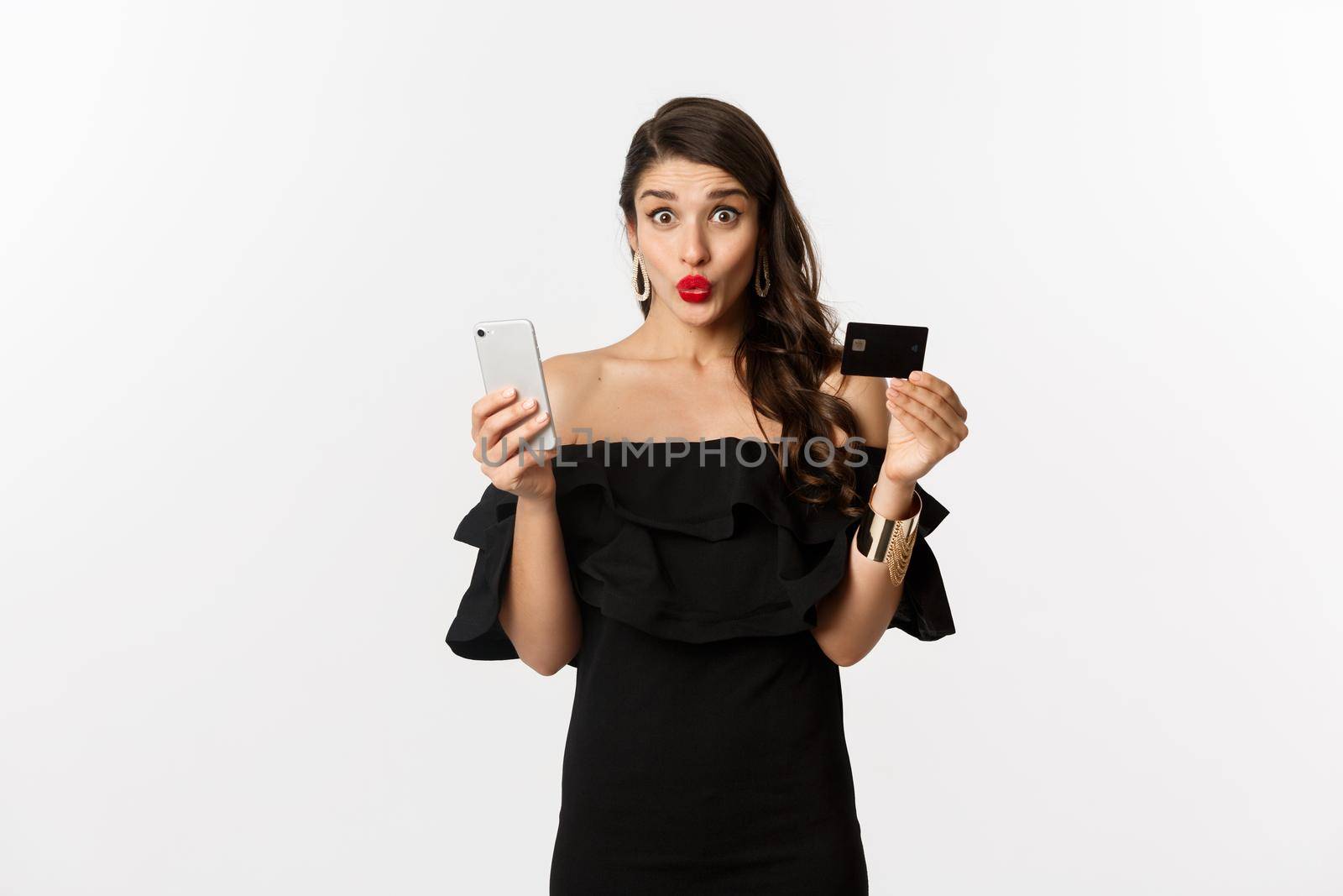 Online shopping concept. Fashionable woman in black dress, holding credit card with smartphone, looking excited, standing over white background.