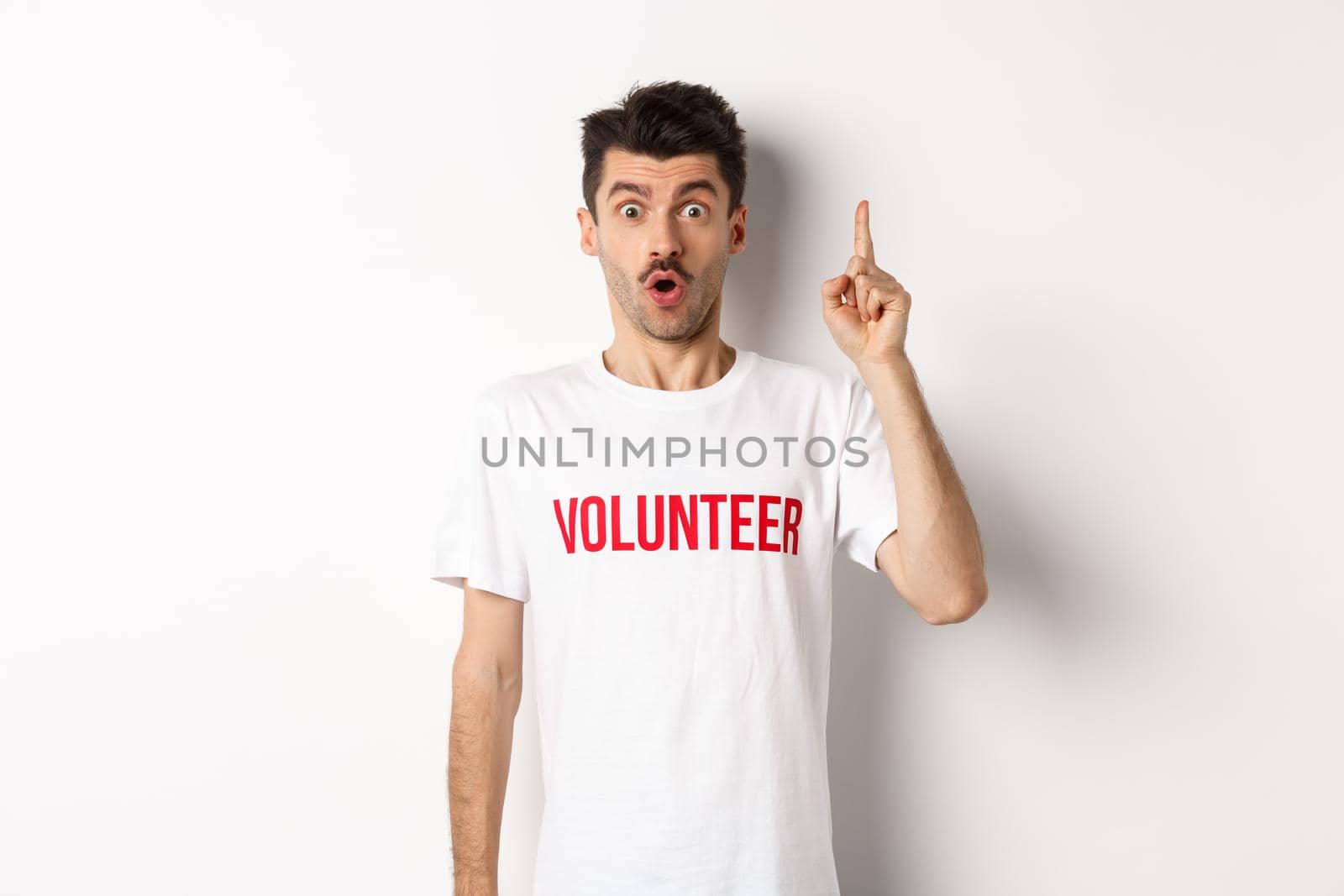 Handsome young man in volunteer t-shirt having an idea, raising finger and saying suggestion, pointing up, standing over white background.