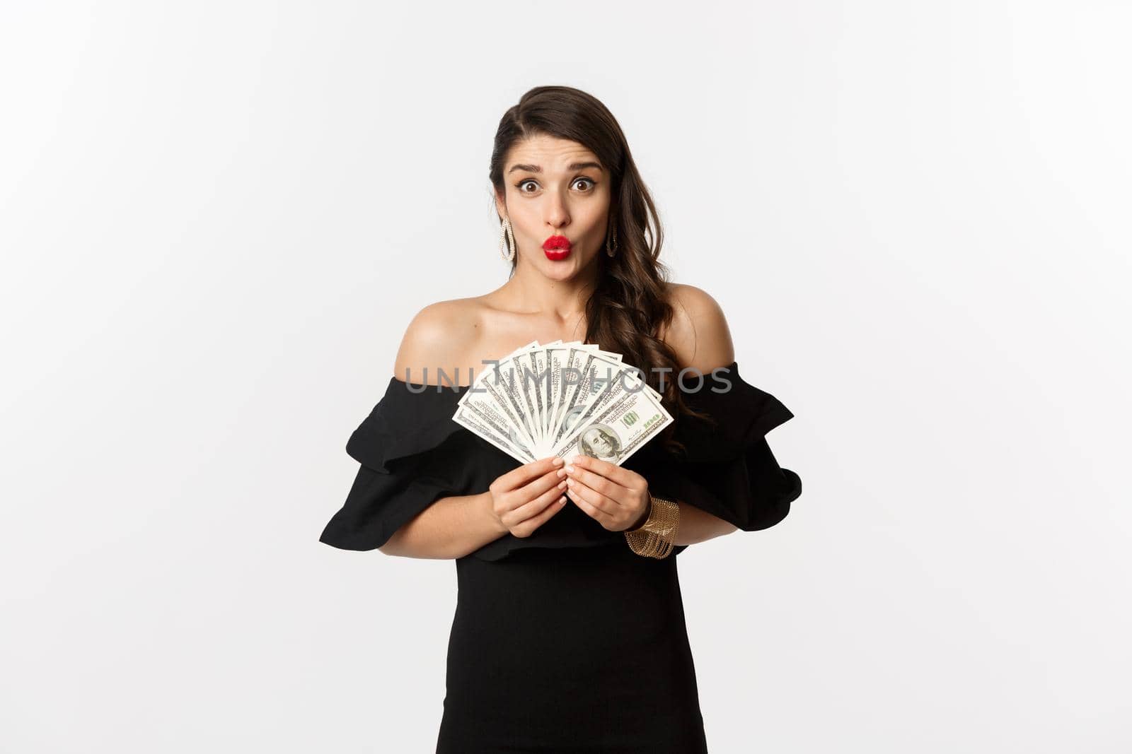 Fashion and shopping concept. Excited woman in black dress, with red lips, showing money dollars and looking amazed at camera, white background.
