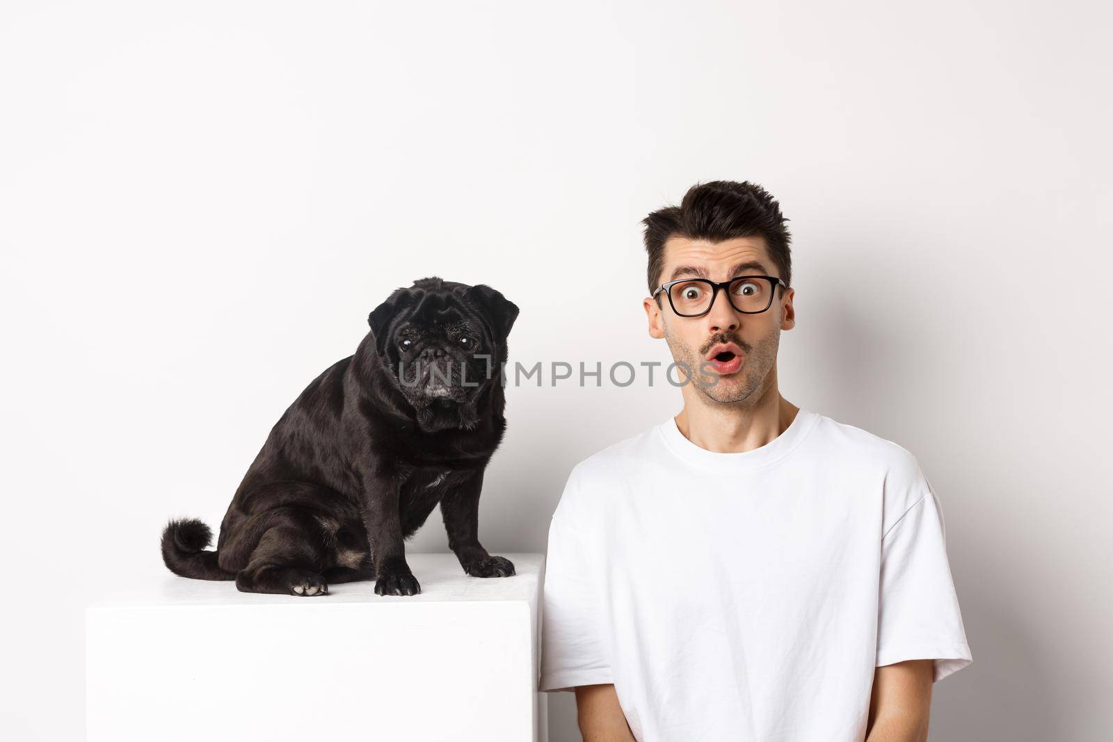 Portrait of dog owner and small cute pug staring at camera surprised and amazed, standing over white background.