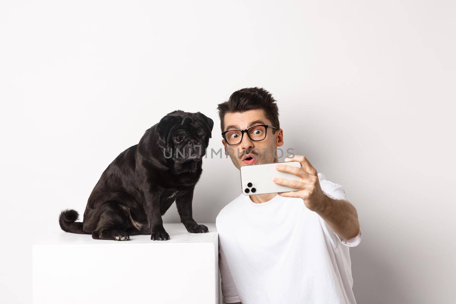 Image of handsome young man taking selfie with cute black dog on smartphone, posing with pug over white background.