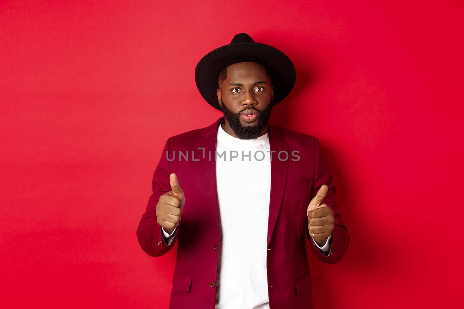 Impressed Black man showing thumbs up and staring amazed at camera, approve and praise something good, red background.