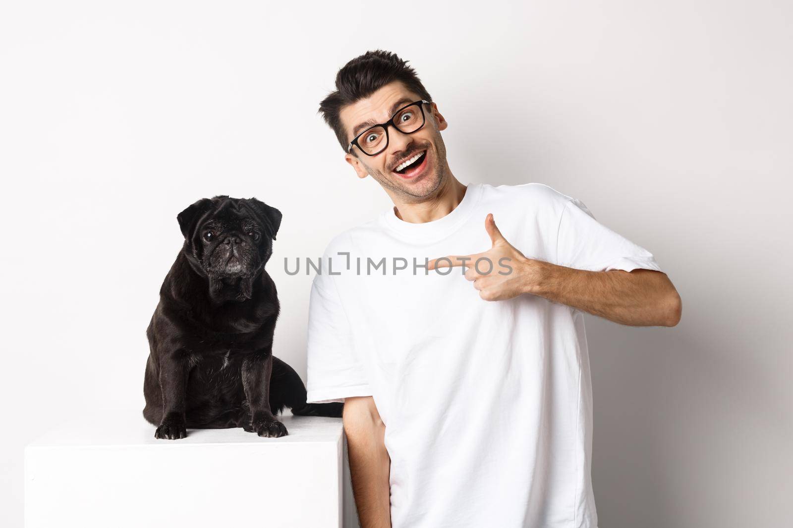 Happy young man showing his cute dog, pointing finger at black pug and smiling, standing over white background.
