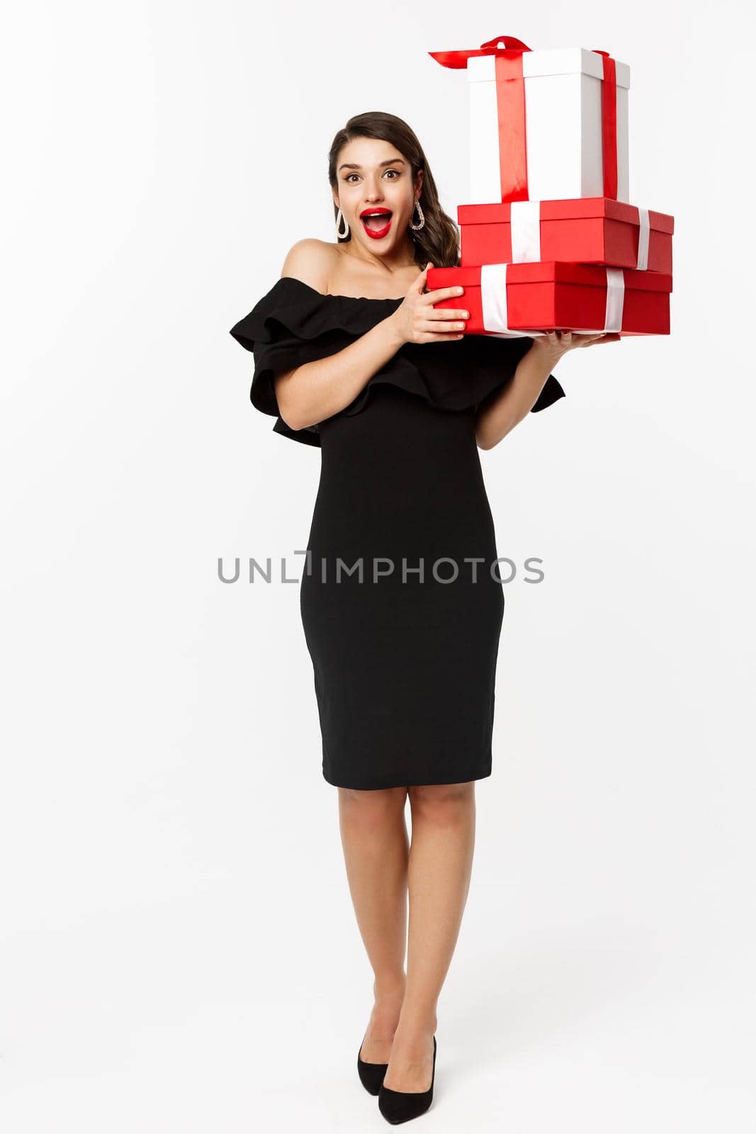 Merry christmas and new year holidays concept. Cheerful lady in black dress holding xmas presents and smiling at camera, standing over white background.