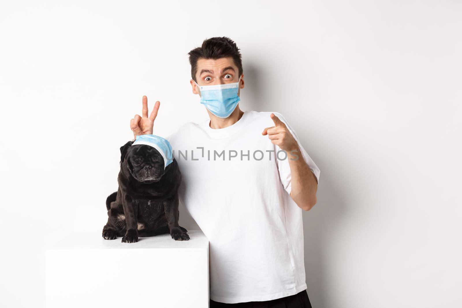 Covid-19, animals and quarantine concept. Happy dog owner and cute pug wearing medical masks, man pointing finger at camera and making funny bunny ears on pet, white background.