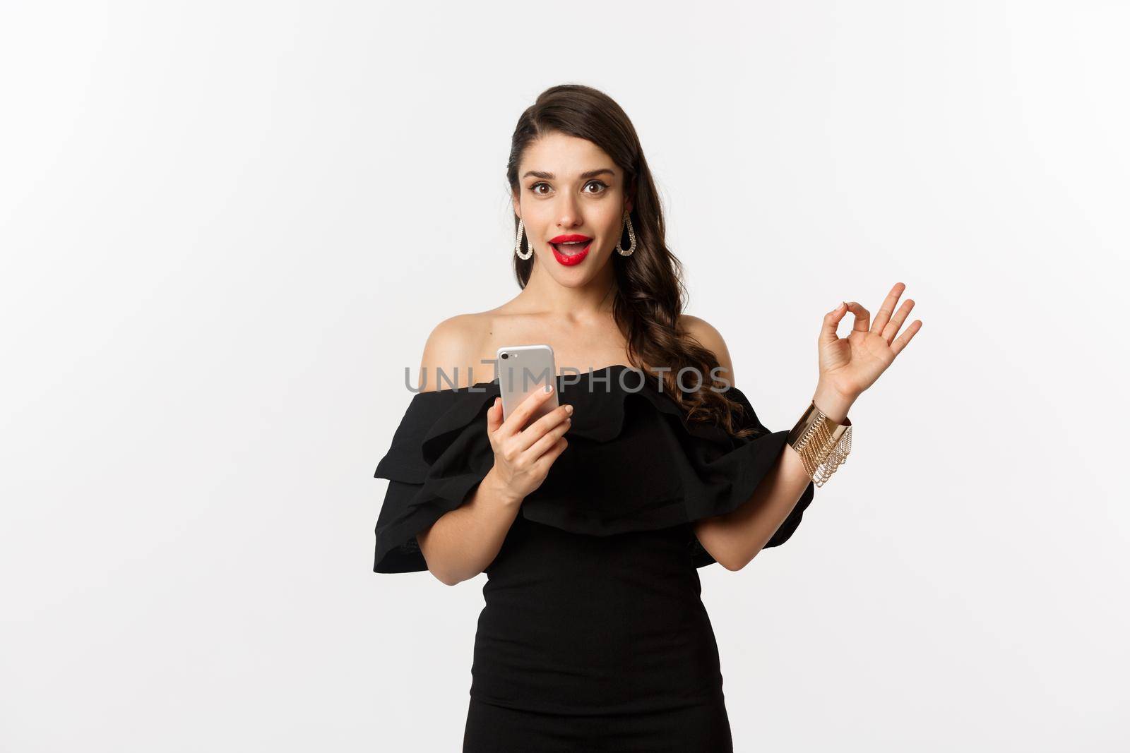 Online shopping concept. Attractive woman in trendy black dress, makeup, showing okay sign in approval and using mobile phone app, white background.