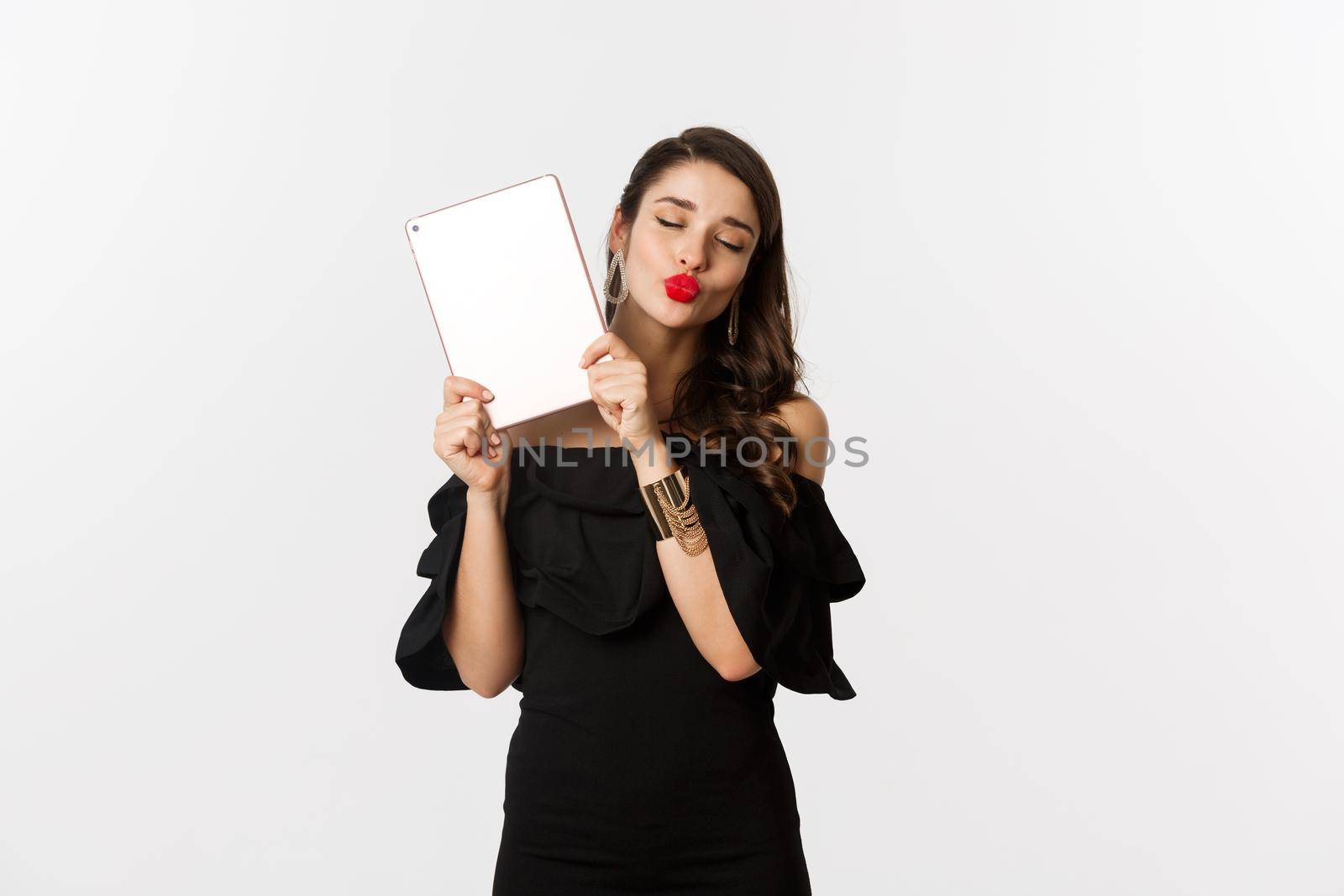 Fashion and shopping concept. Beautiful woman with red lips, wearing black dress, showing digital tablet and making kissing face, white background.