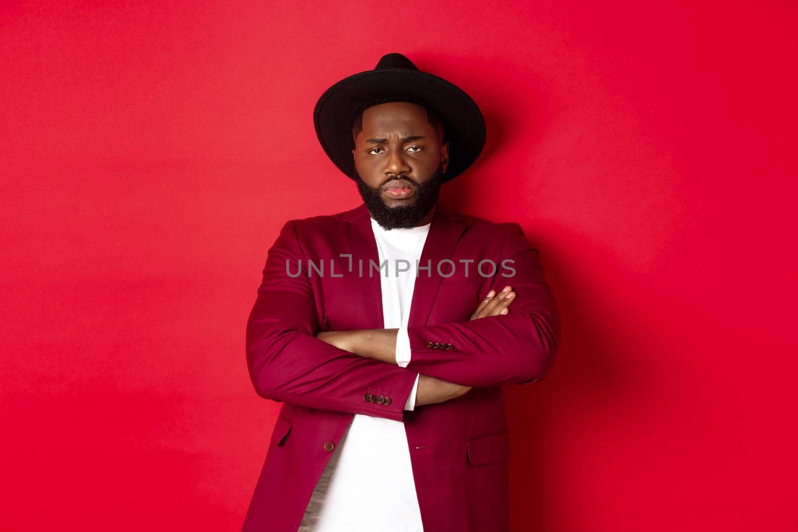 Reluctant and upset Black man waiting for apology, cross arms on chest and looking offended at camera, red background by Benzoix