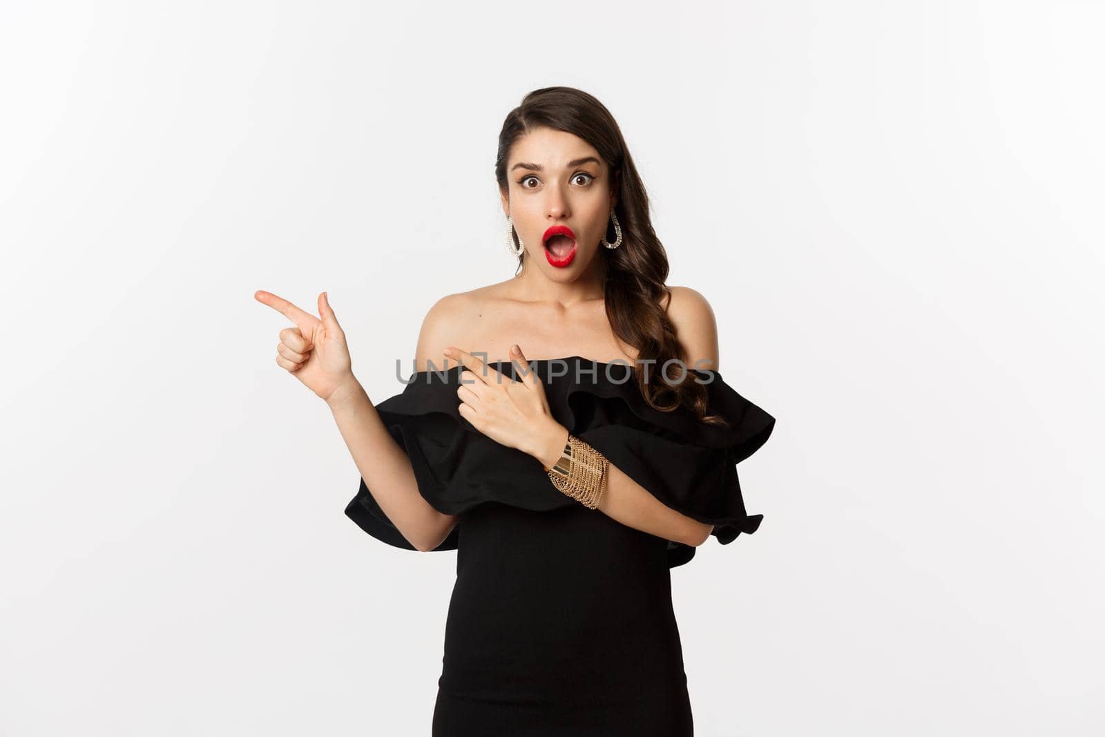 Fashion and beauty. Surprised woman in black glamour dress pointing fingers left, showing advertisement and staring amazed, white background by Benzoix