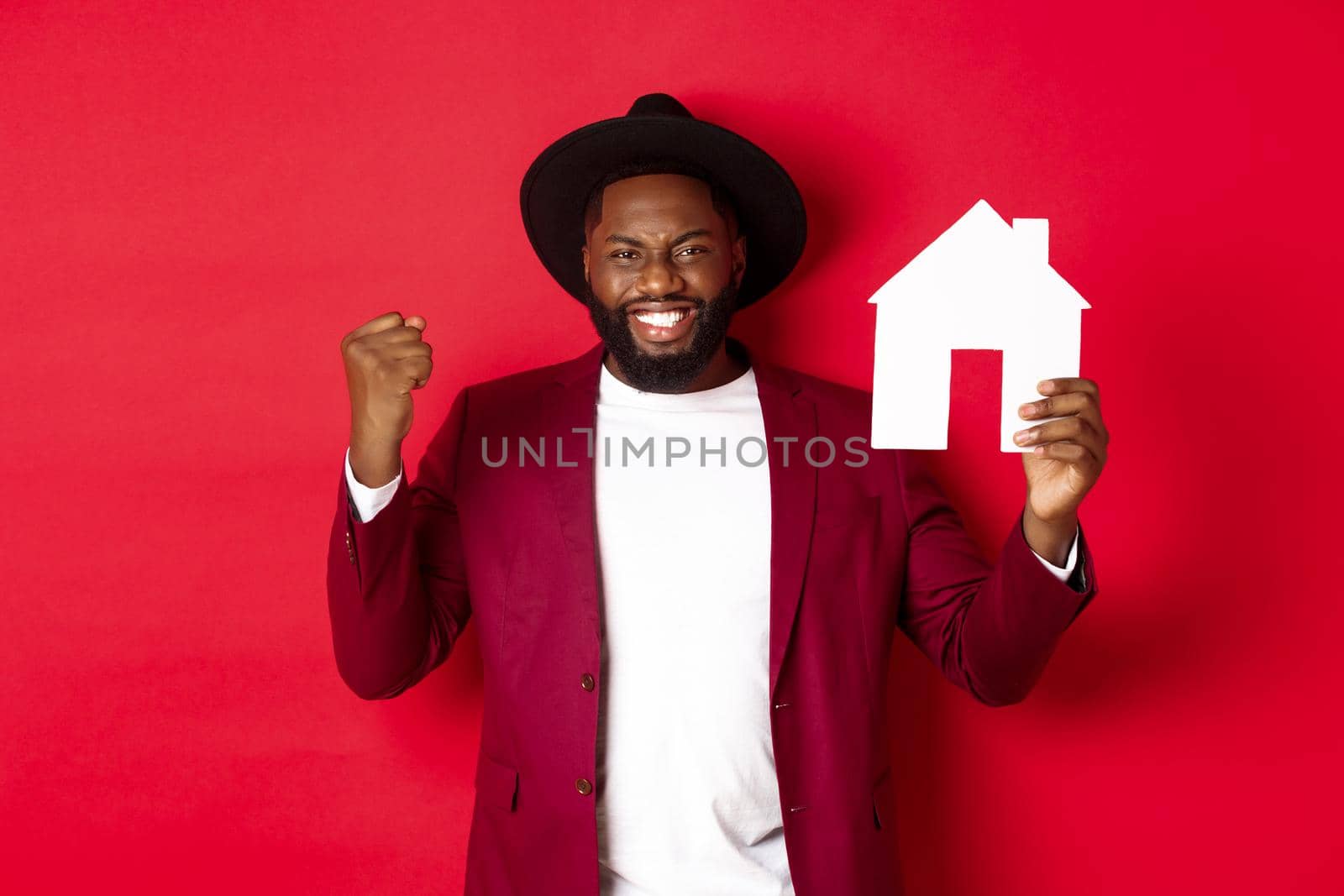 Real estate. Cheerful Black man rejoicing and showing paper home maket, standing over red background by Benzoix