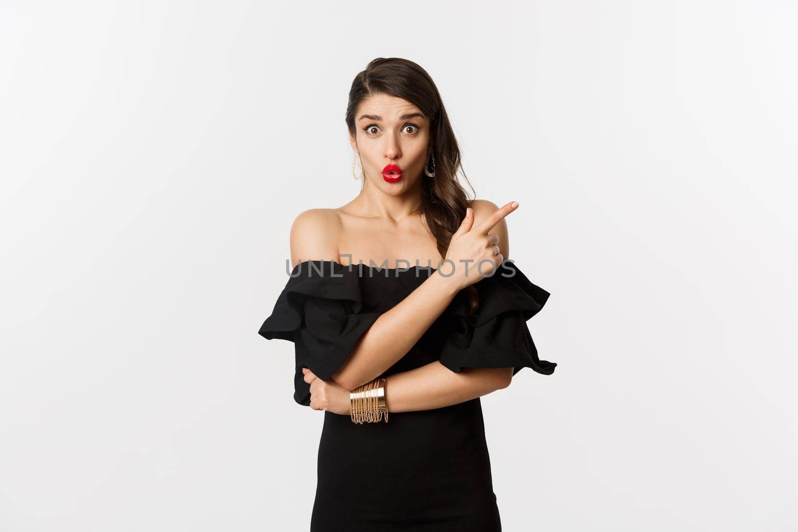 Fashion and beauty. Surprised woman in black dress pointing finger at upper right corner, showing advertisement, standing over white background.