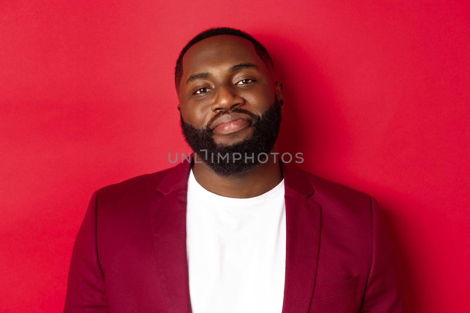 Close-up of handsome bearded plump guy smiling at camera shy, standing in stylish blazer on t-shirt, standing over red background.