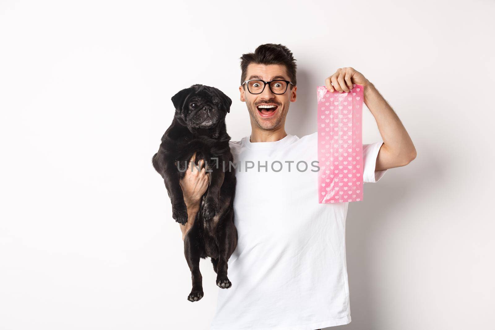 Image of hipster guy pet owner, holding cute black pug and dog poop bag, standing over white background.