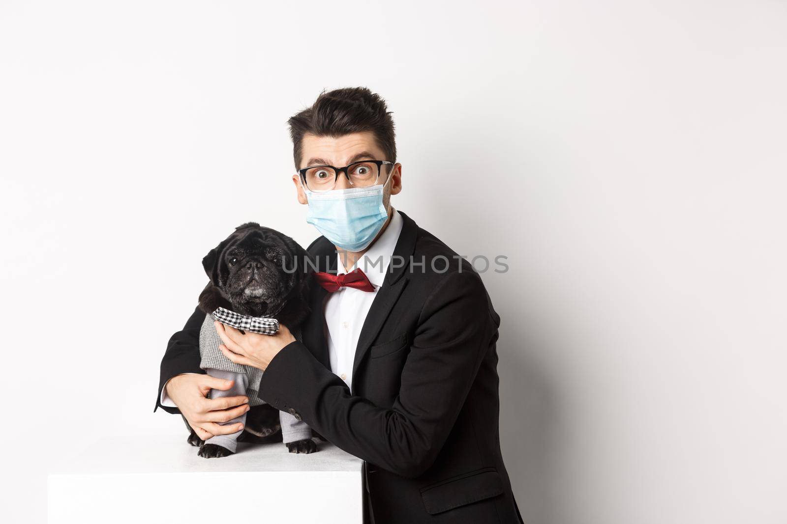 Coronavirus, pets and celebration concept. Happy dog owner in suit and face mask hugging cute black pug in costume, standing over white background.