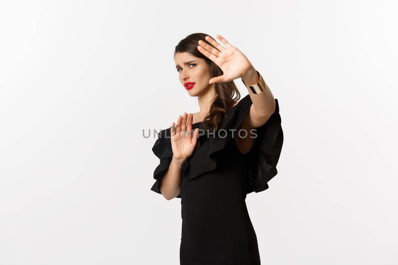 Fashion and beauty. Reluctant and worried woman asking to stay away, showing stop gesture and looking scared, standing in black dress over white background.