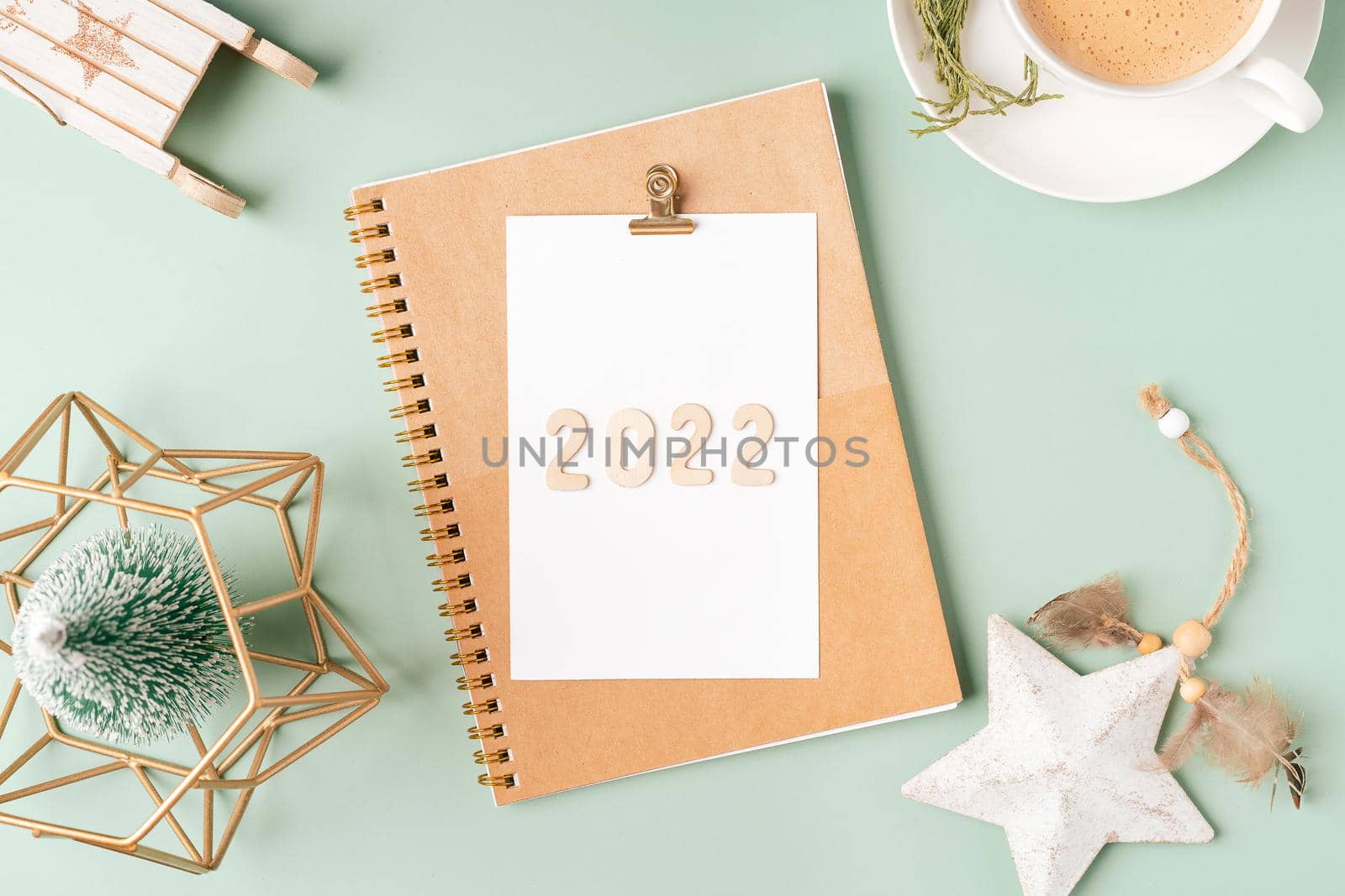 Top view Desktop Christmas notepad with 2022 text. Flat lay on green mint table background with planner, cup of coffee, tree brunch, Christmas eco nature decoration, notebook and stationery by Ostanina