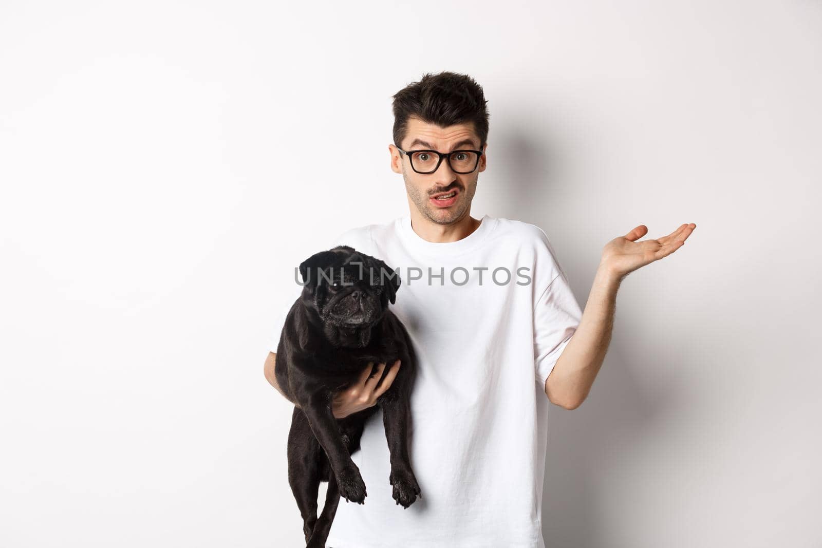 Image of handsome young man holding black pug and looking confused. Guy shrugging shoulders and staring indecisive at camera, carry dog in arm, white background.