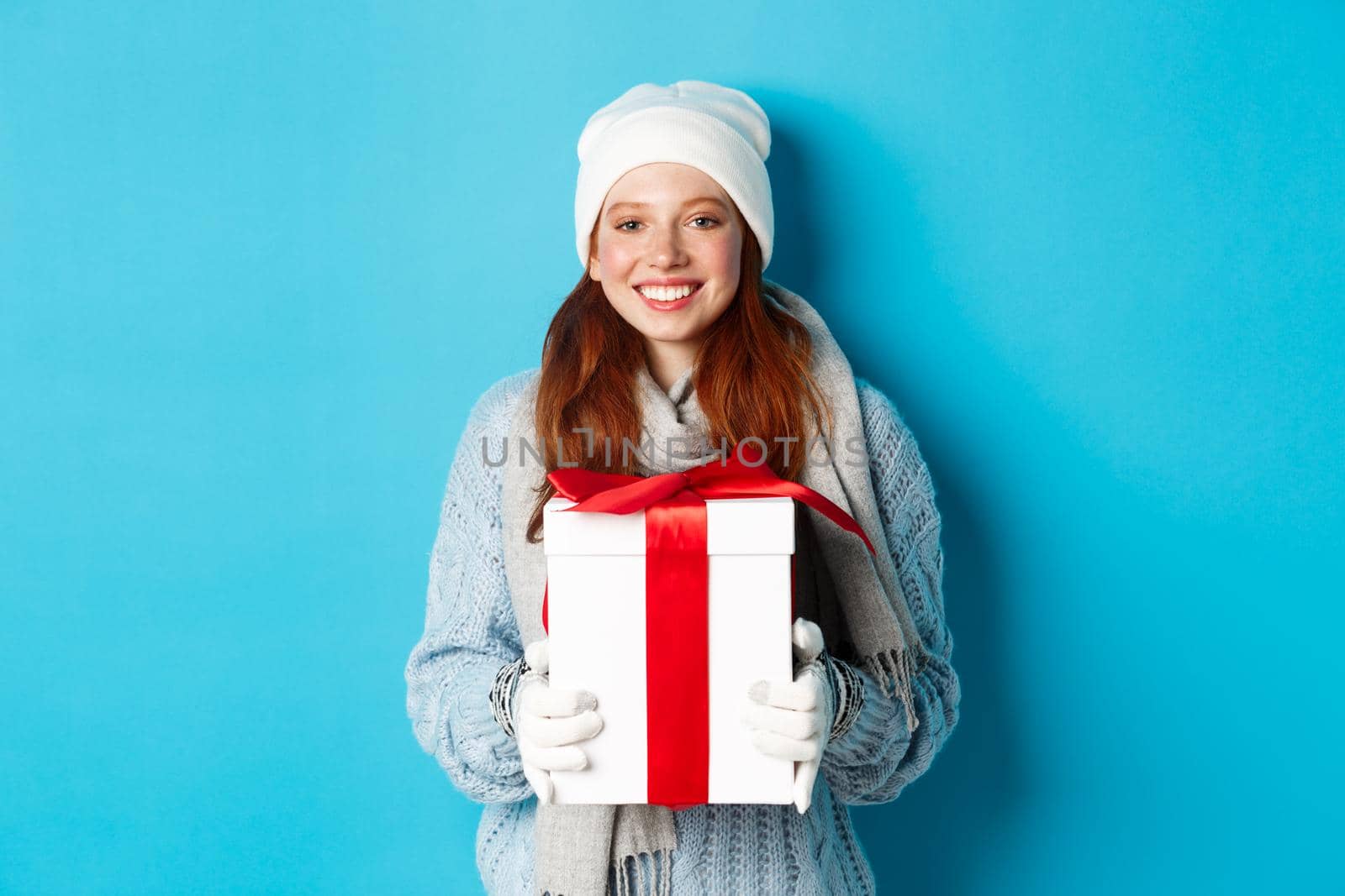 Winter holidays and Christmas sales concept. Cute redhead teeanage girl in beanie, sewater and scarf holding gift in wrapped box, smiling, wishing merry xmas, standing over blue background.