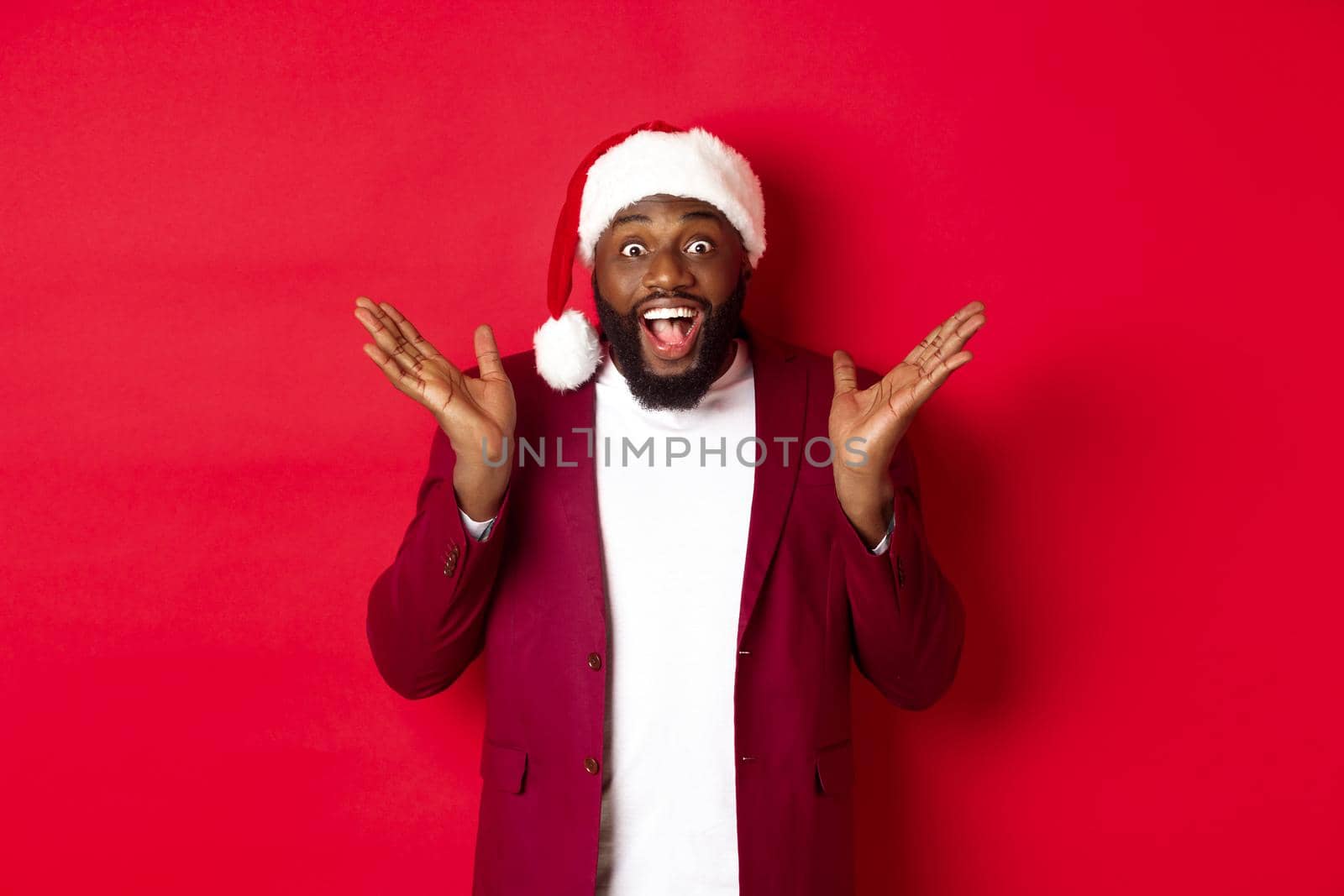 Christmas, party and holidays concept. Surprised and happy african american man in santa hat, looking with rejoice at camera, standing against red background by Benzoix