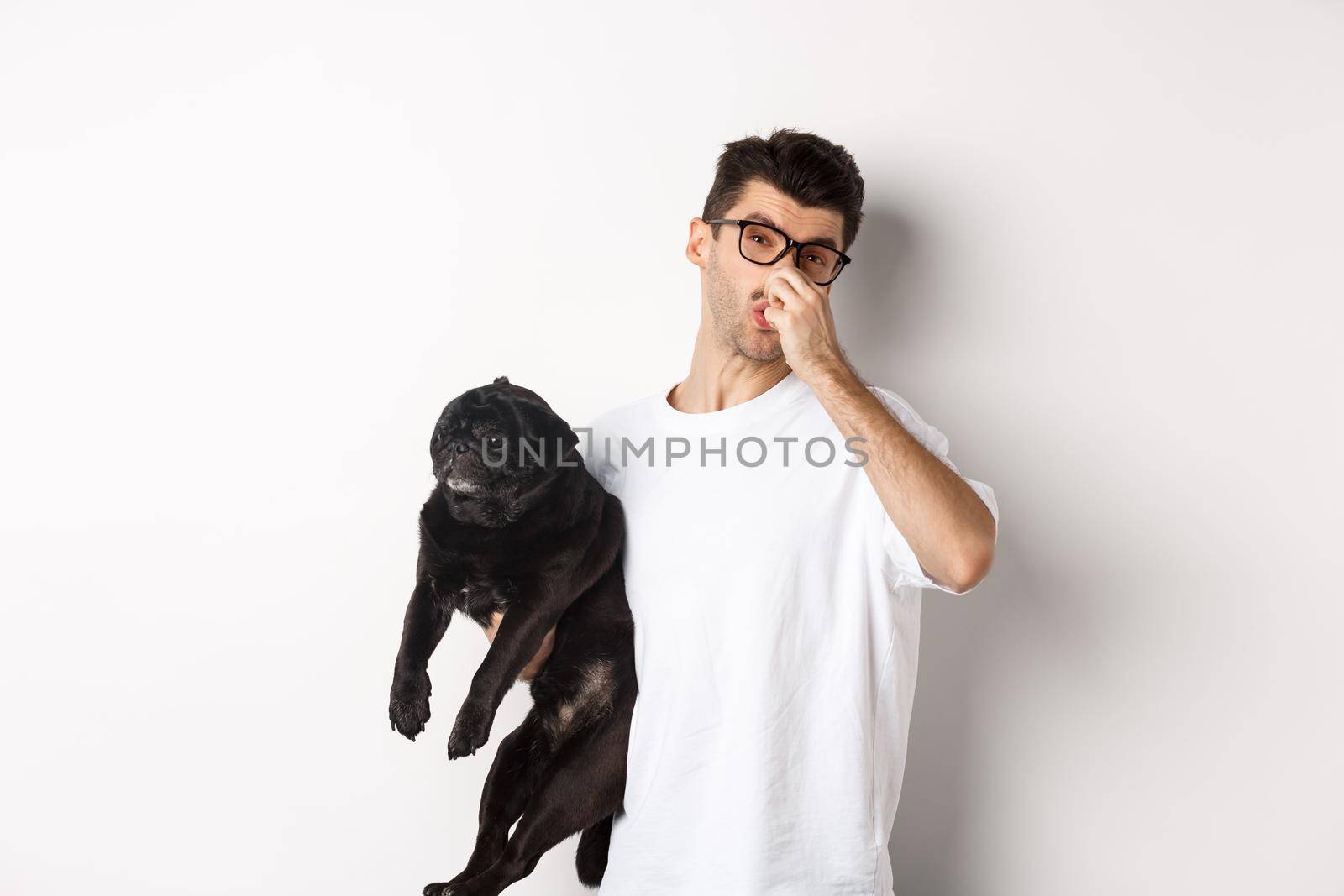 Young man shut nose as holding pug, disgusted with bad smell fart of animal, standing over white background.